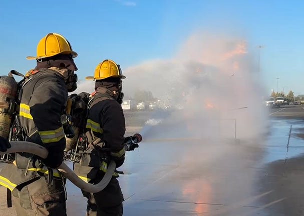 Firefighters in yellow helmets extinguishing a fire with a hose, surrounded by smoke and water spray.