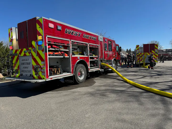A red fire truck labeled "West Metro Fire Rescue" with firefighters and equipment, set against a clear blue sky.