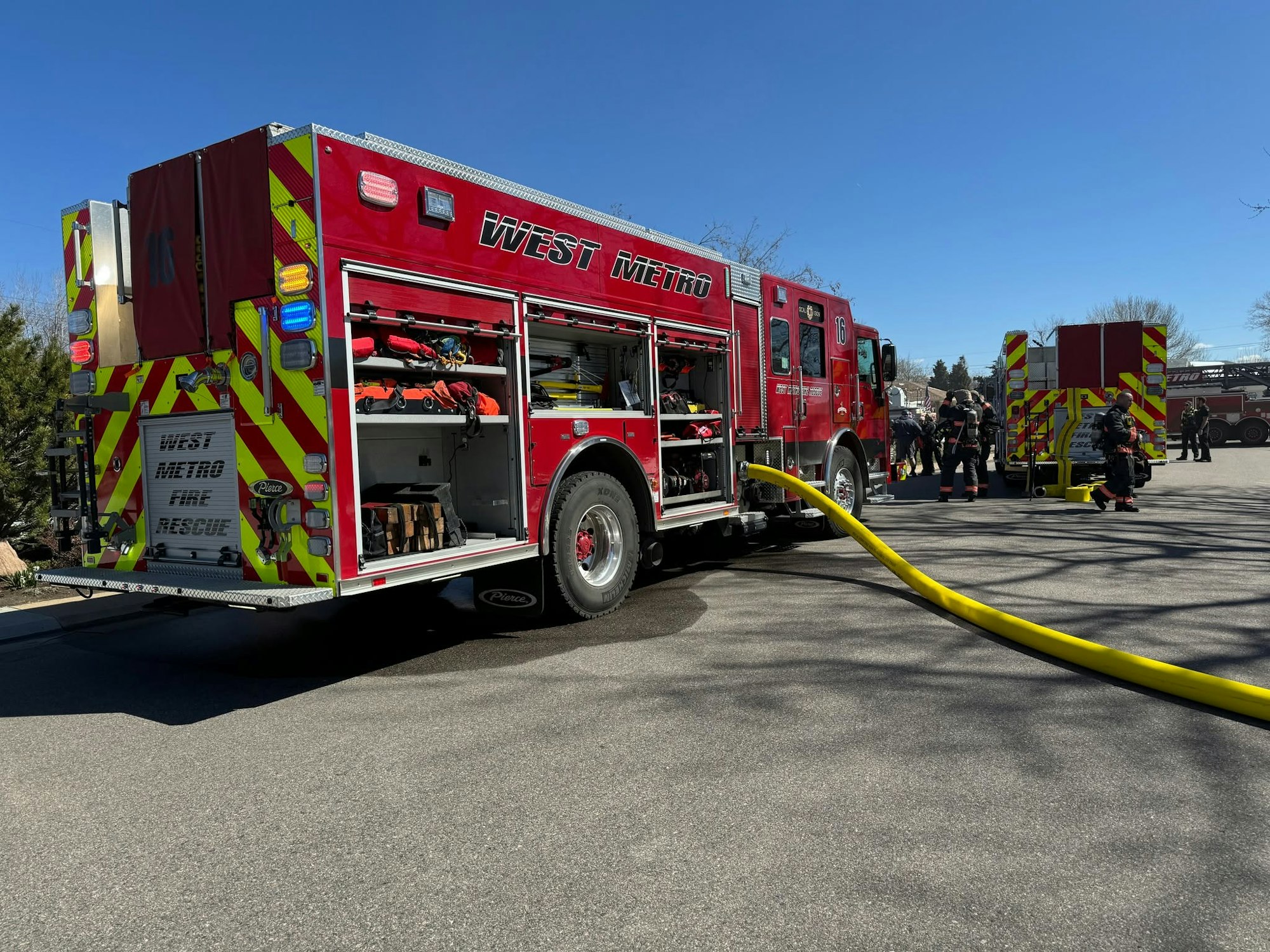 A red fire truck labeled "West Metro Fire Rescue" with firefighters and equipment, set against a clear blue sky.