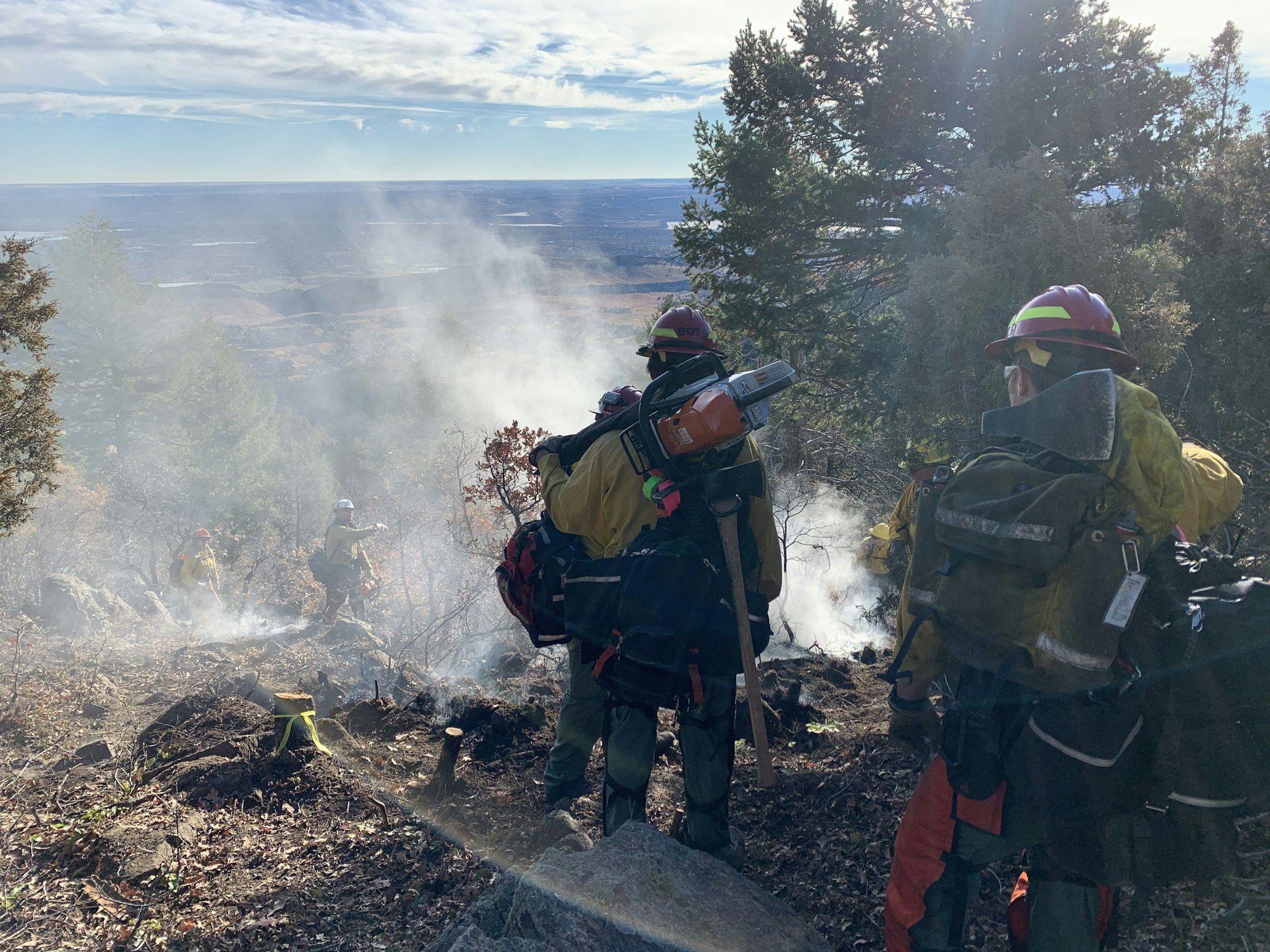 West Metro firefighters working on mop up in a burned area following a wild land fire. All firefighters are carrying line packs, one is carrying a chain saw. They are standing in a forested area near a rock outcropping. You can see smoke in front of them.