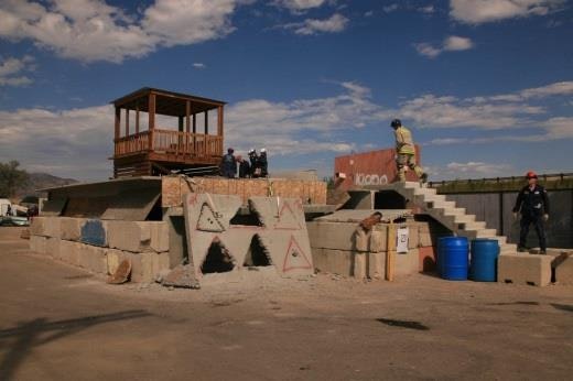 Concrete obstacle course with people, ramps, a wooden shelter, and barrels under a blue sky.
