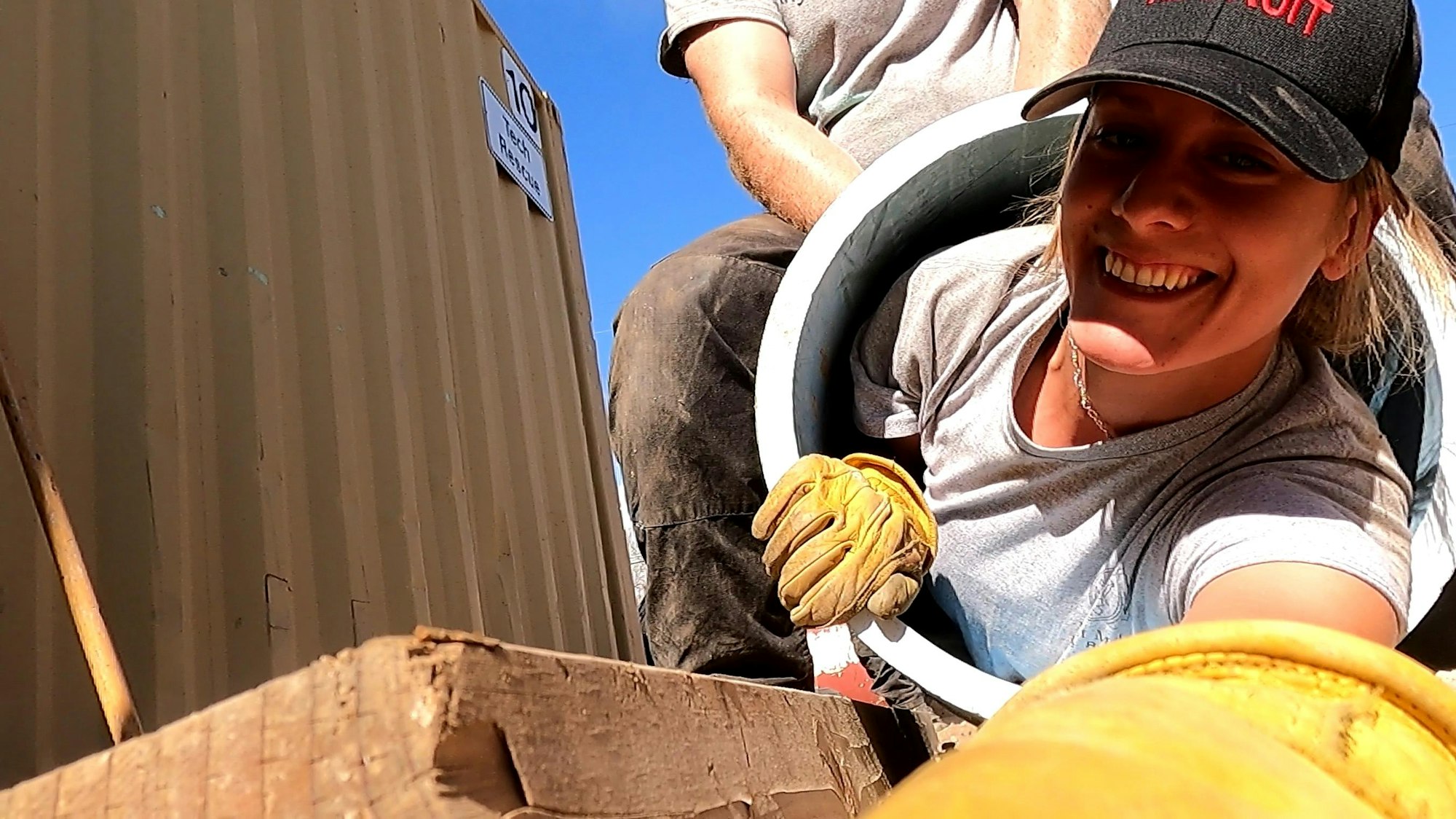 Two people working, one smiling at the camera, wearing gloves and a cap, with a large tube and a wooden structure in the background.