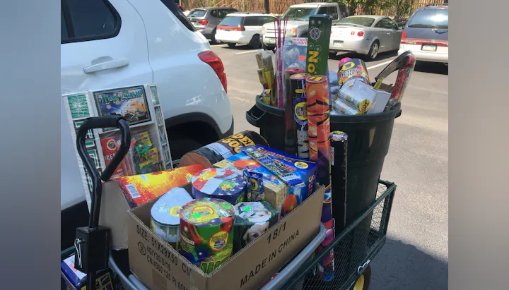 A cart filled with various types of fireworks, including rockets, sparklers, and colorful explosive items, parked in a lot.