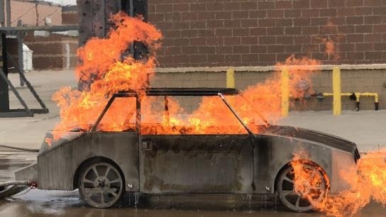 A metal car model engulfed in flames outdoors, likely a controlled fire scenario.
