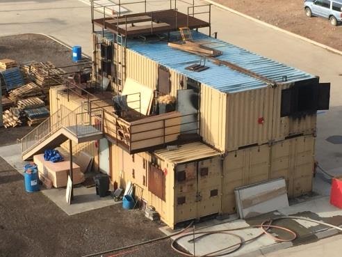A makeshift building made from shipping containers, with a staircase and scaffolding on top, surrounded by construction materials.