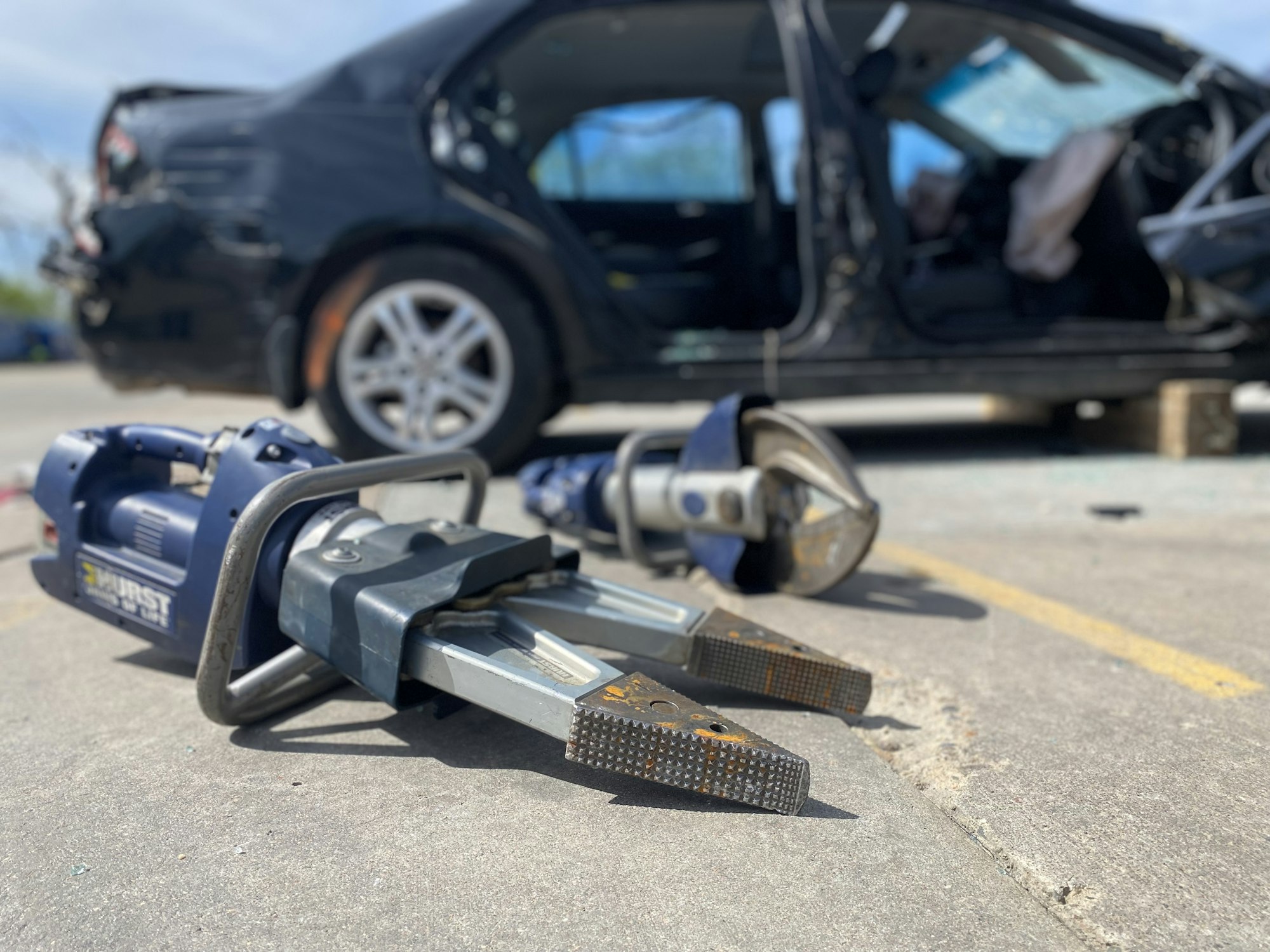 Jaws of Life rescue tool in focus with a damaged car in the background.
