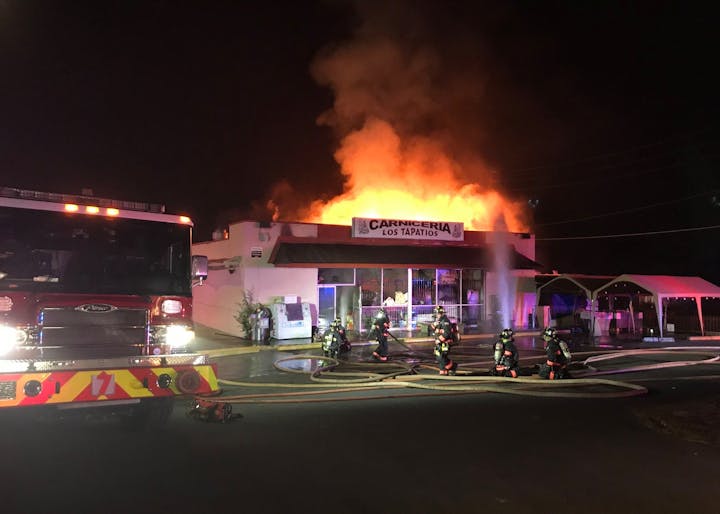 Firefighters respond to a large fire at "Carniceria Los Tapatios" with flames visible and fire hoses in use.