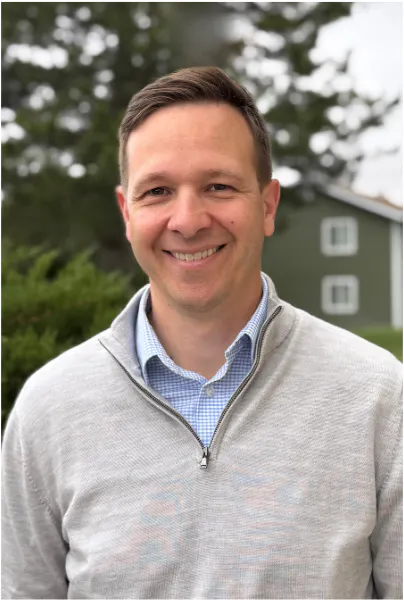 A smiling man in a light sweater poses outdoors, with greenery and a building in the background.