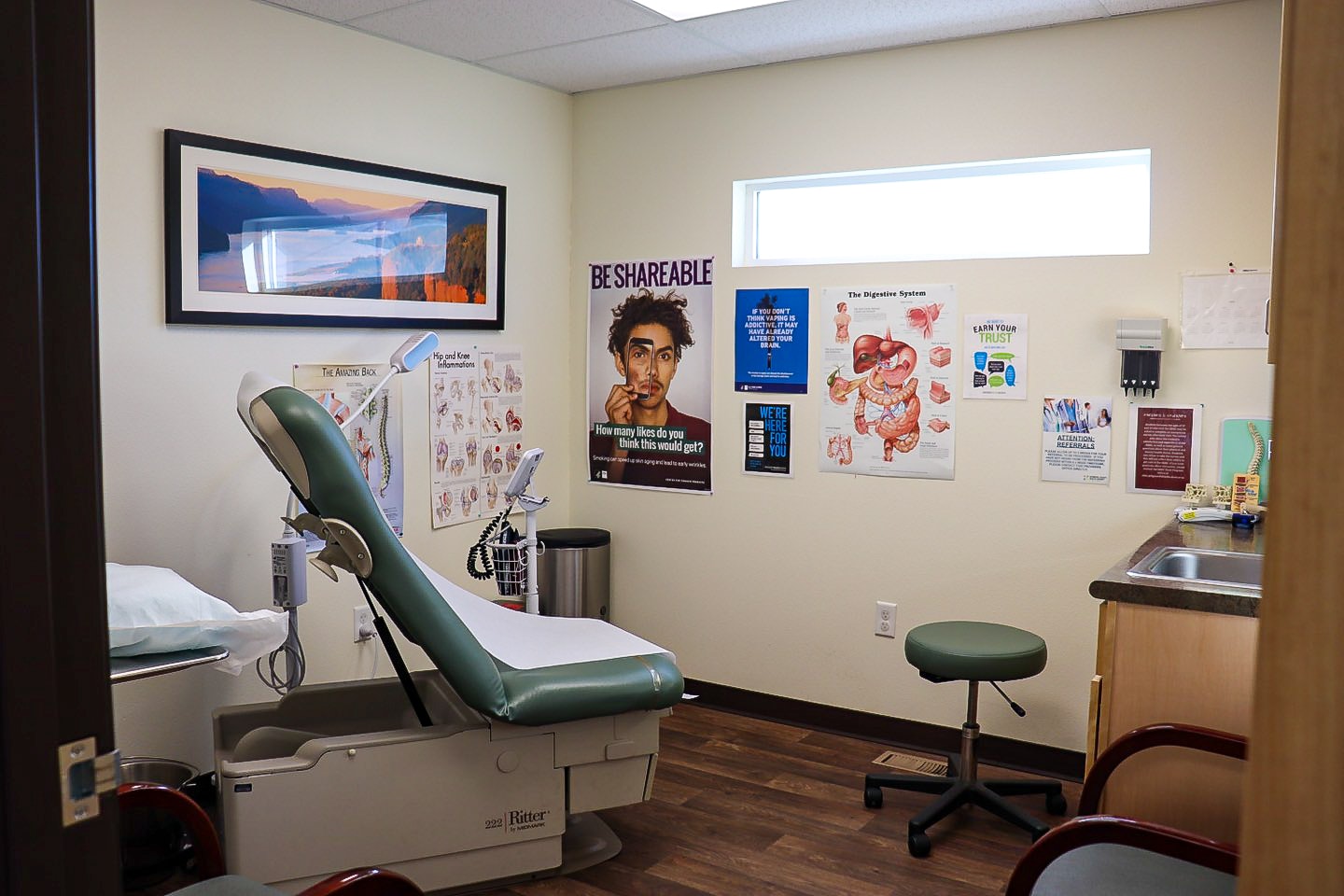 A medical examination room with a patient chair, sink, educational posters, and a landscape painting on the wall.