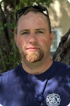 A man with short hair and a goatee, wearing a blue shirt, stands in front of a tree, looking directly at the camera.