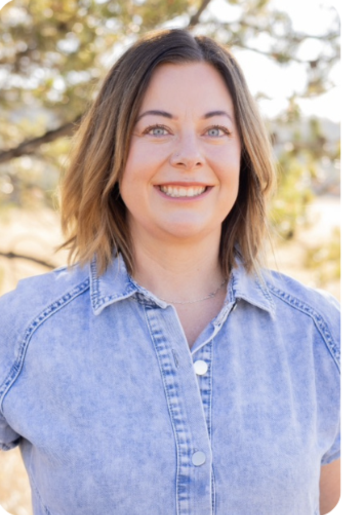 A smiling woman wearing a denim shirt with a blurred natural backdrop.