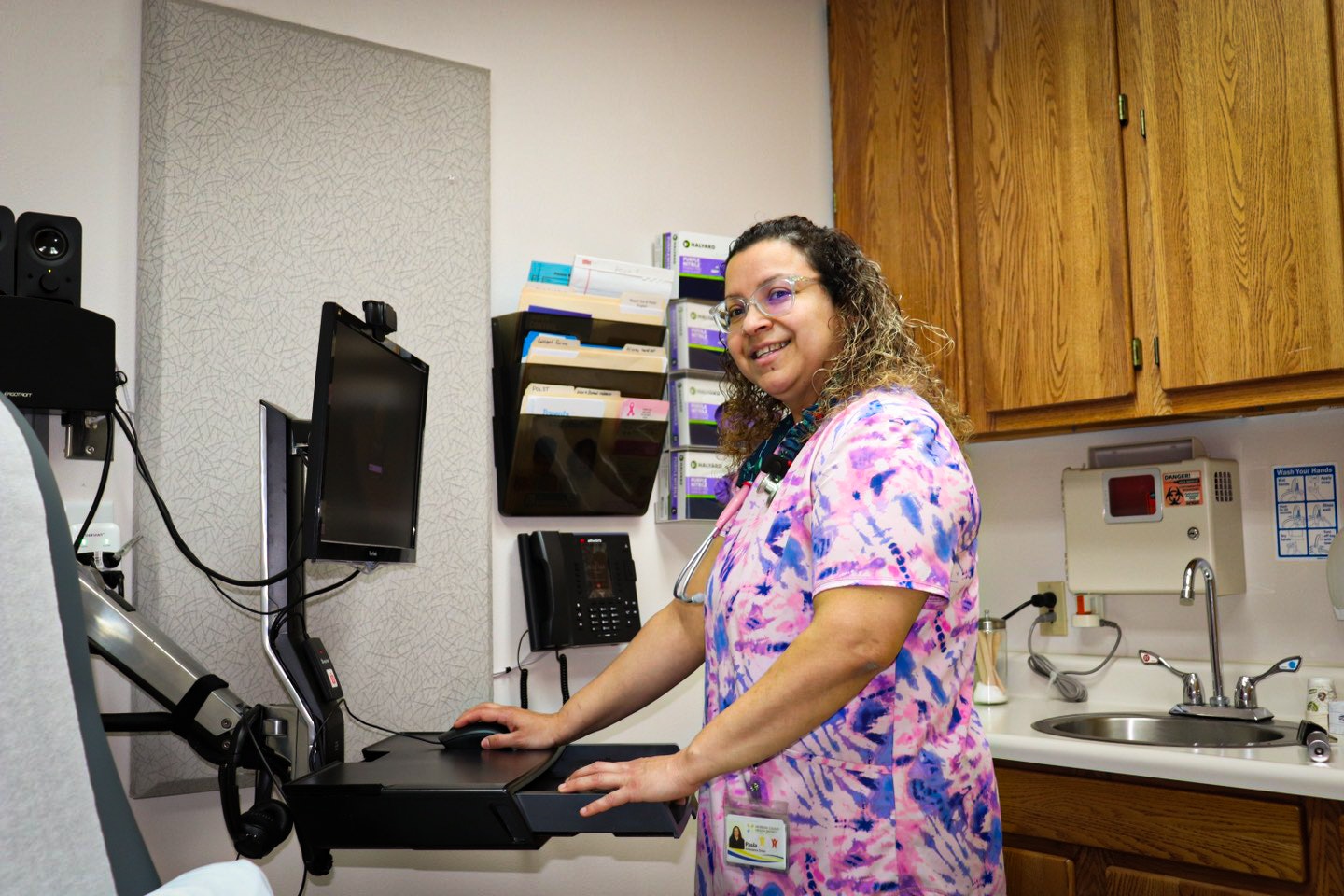 A smiling healthcare professional in scrubs at a computer workstation in a medical office.