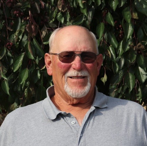An older man with sunglasses and a polo shirt smiles in front of a leafy background.