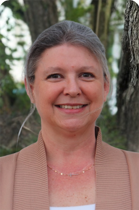 A smiling woman with gray hair is wearing a light brown cardigan and a delicate necklace, posed outdoors near trees.