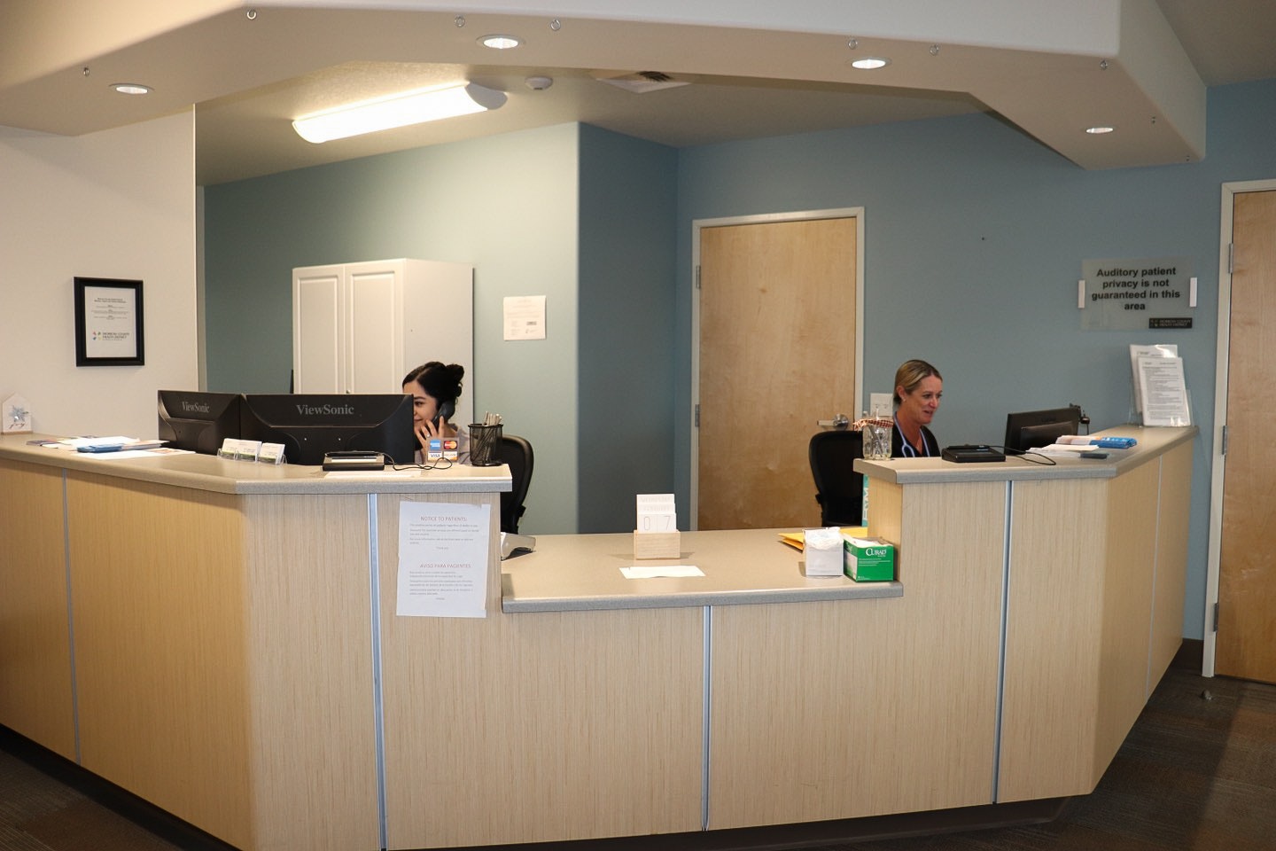 A reception area with two people working behind the desk, computers, signs, and office supplies.