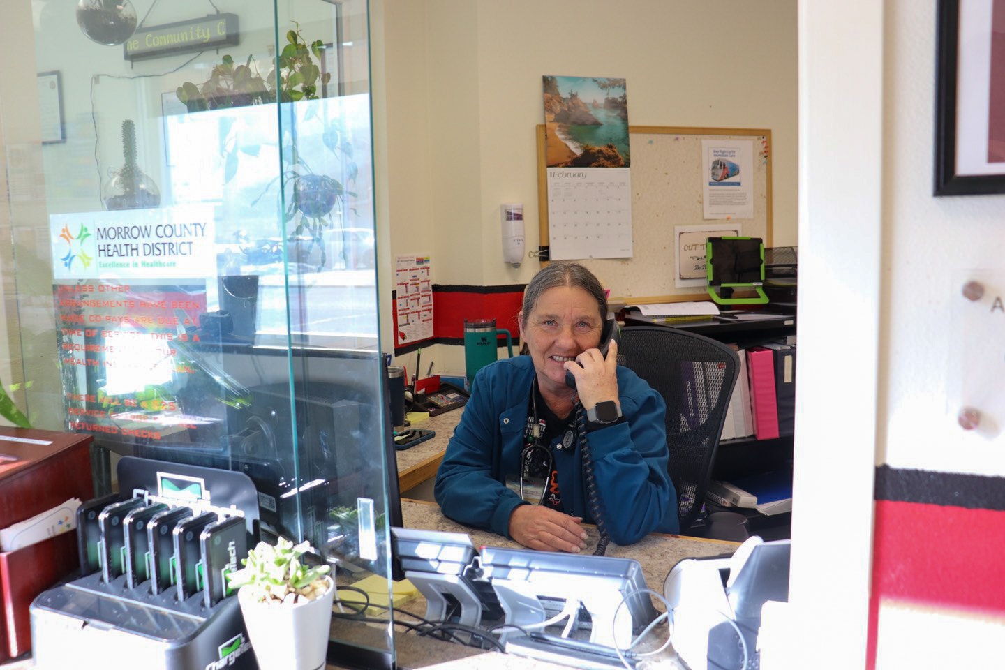 A smiling person at a desk with a phone, computer monitors, and signage for Morrow County Health District.