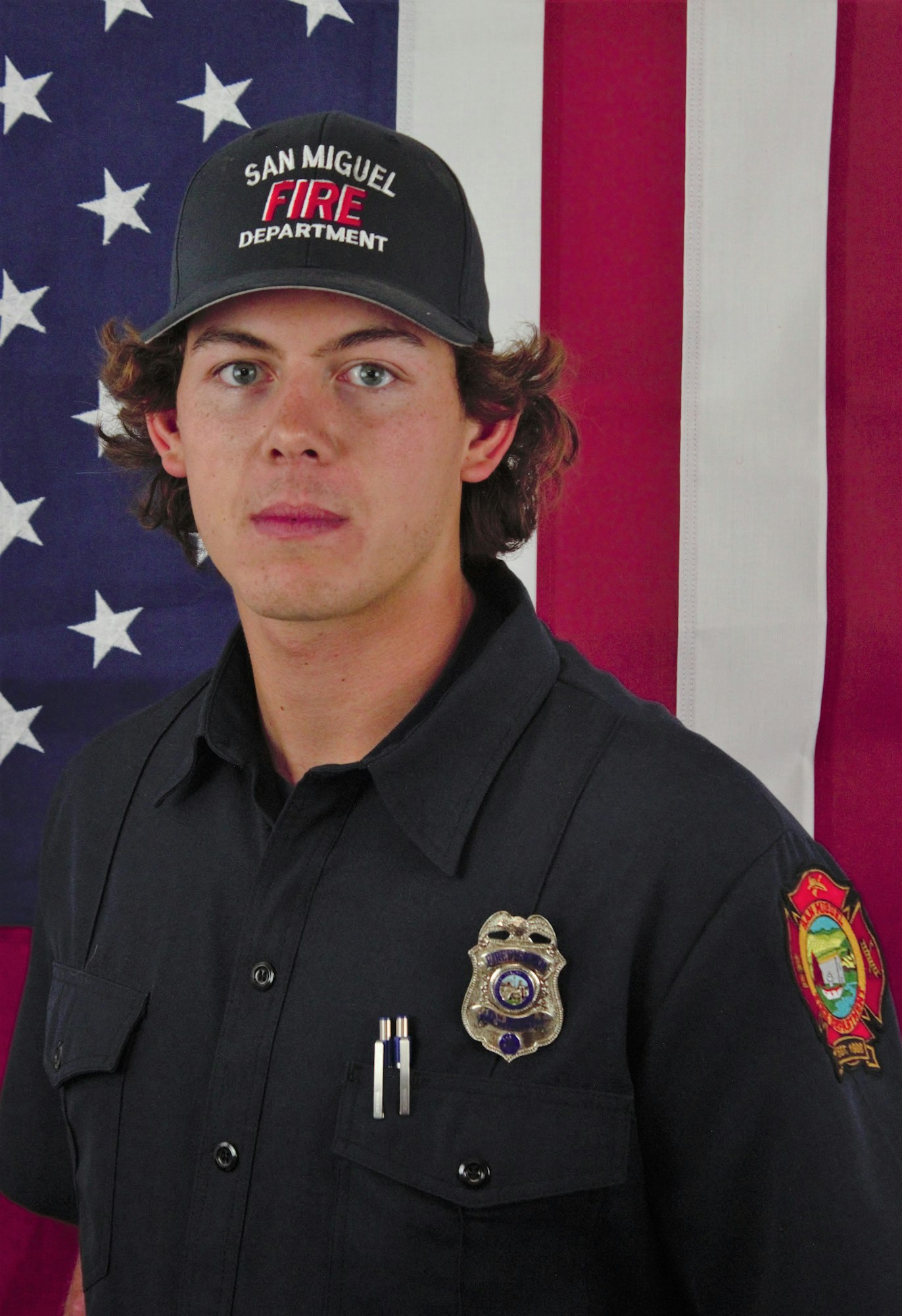 A person in uniform with a "San Miguel Fire Department" cap, standing in front of a U.S. flag.