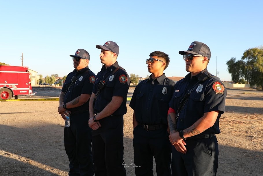Four firefighters in uniform standing in a line outdoors with a fire truck in the background.