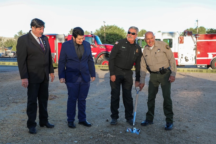 Four men in formal attire standing in front of emergency vehicles, with one man holding a ceremonial shovel.