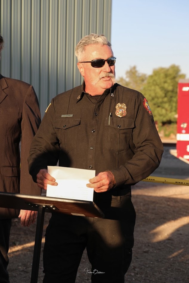 Person in uniform with badge and sunglasses, holding a paper, partial view of another person, outdoor setting, sunny.