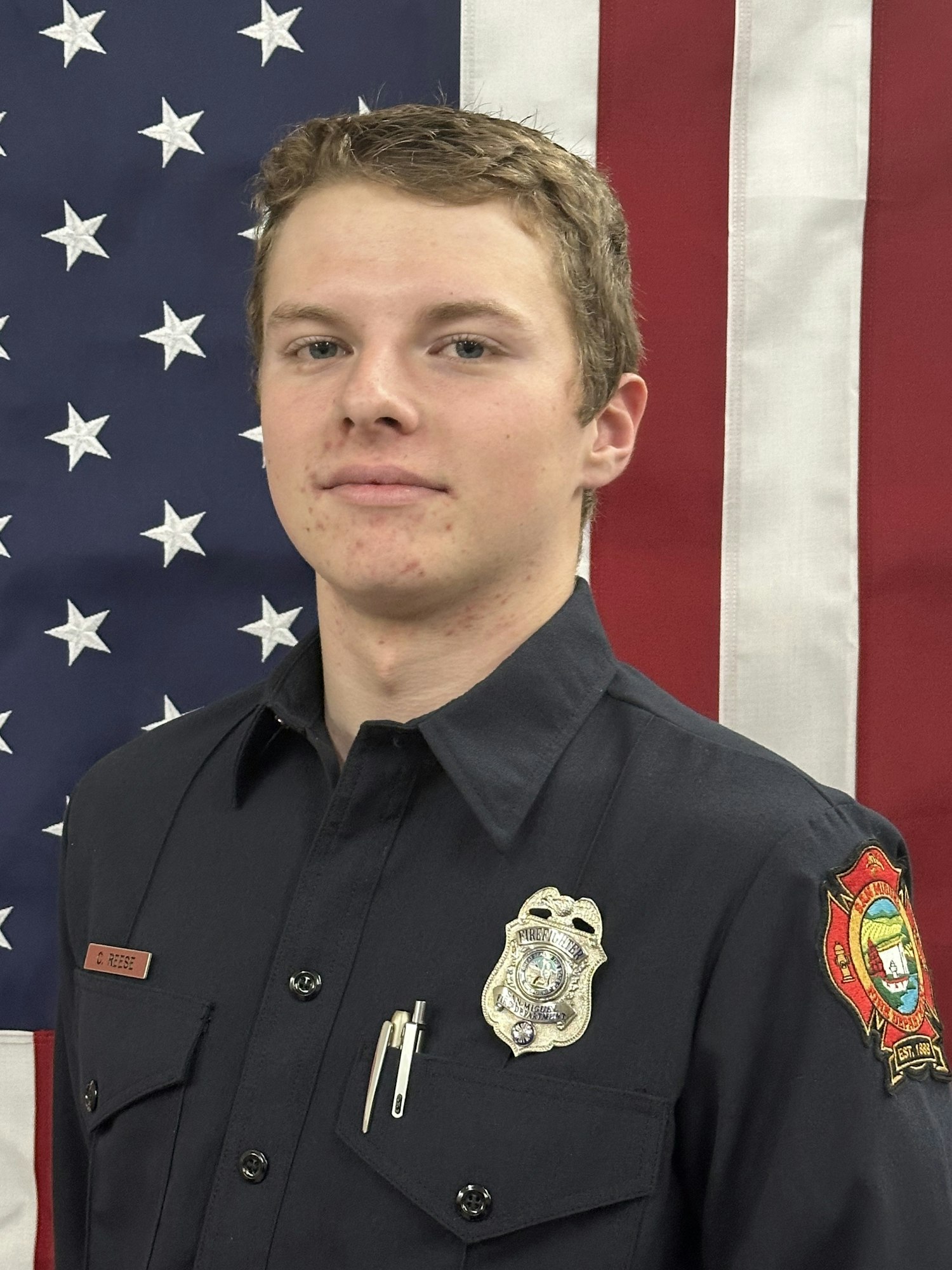 A person in a uniform with badges and pens, standing in front of a U.S. flag.