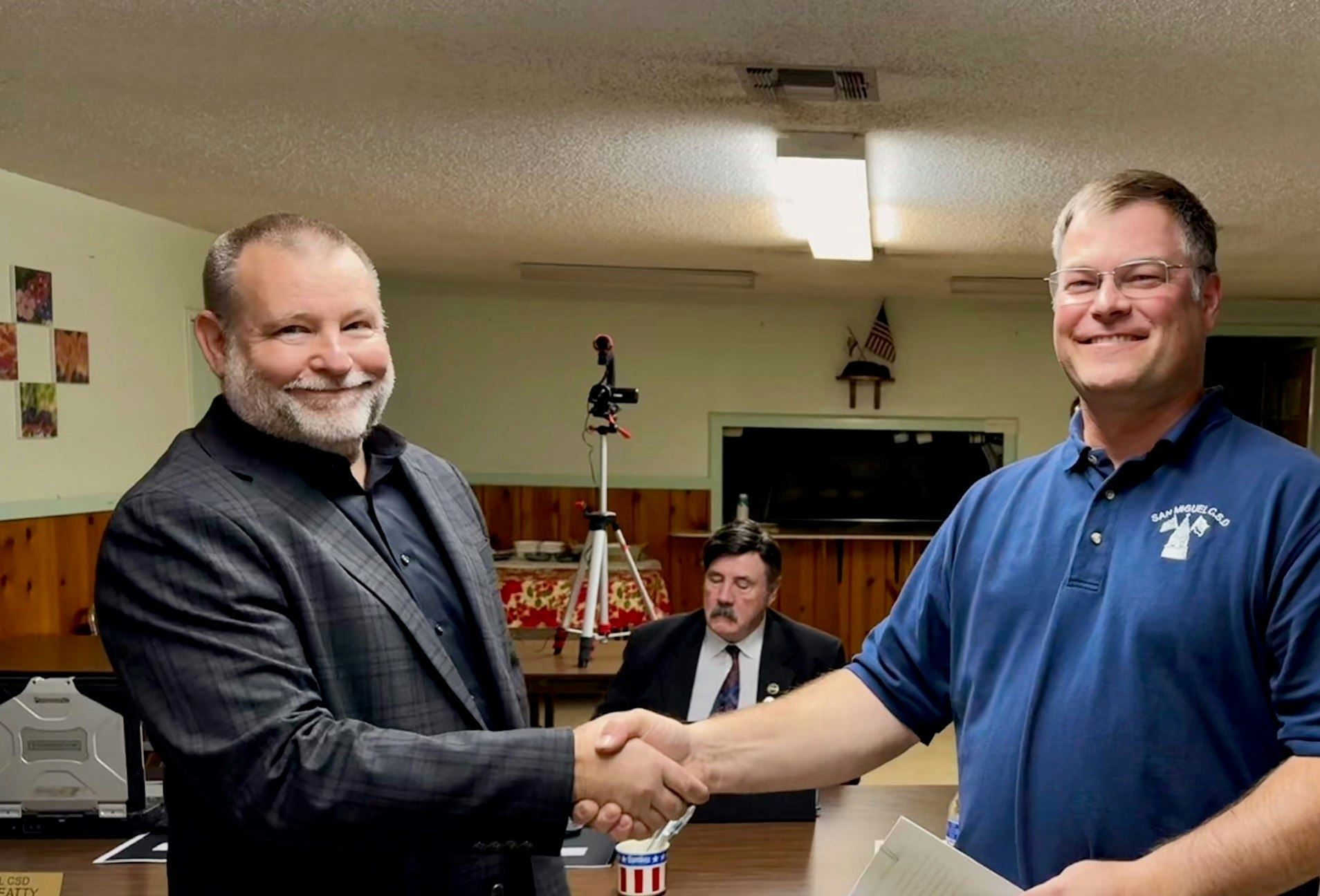 Two men are shaking hands in a room, while another man sits in the background.