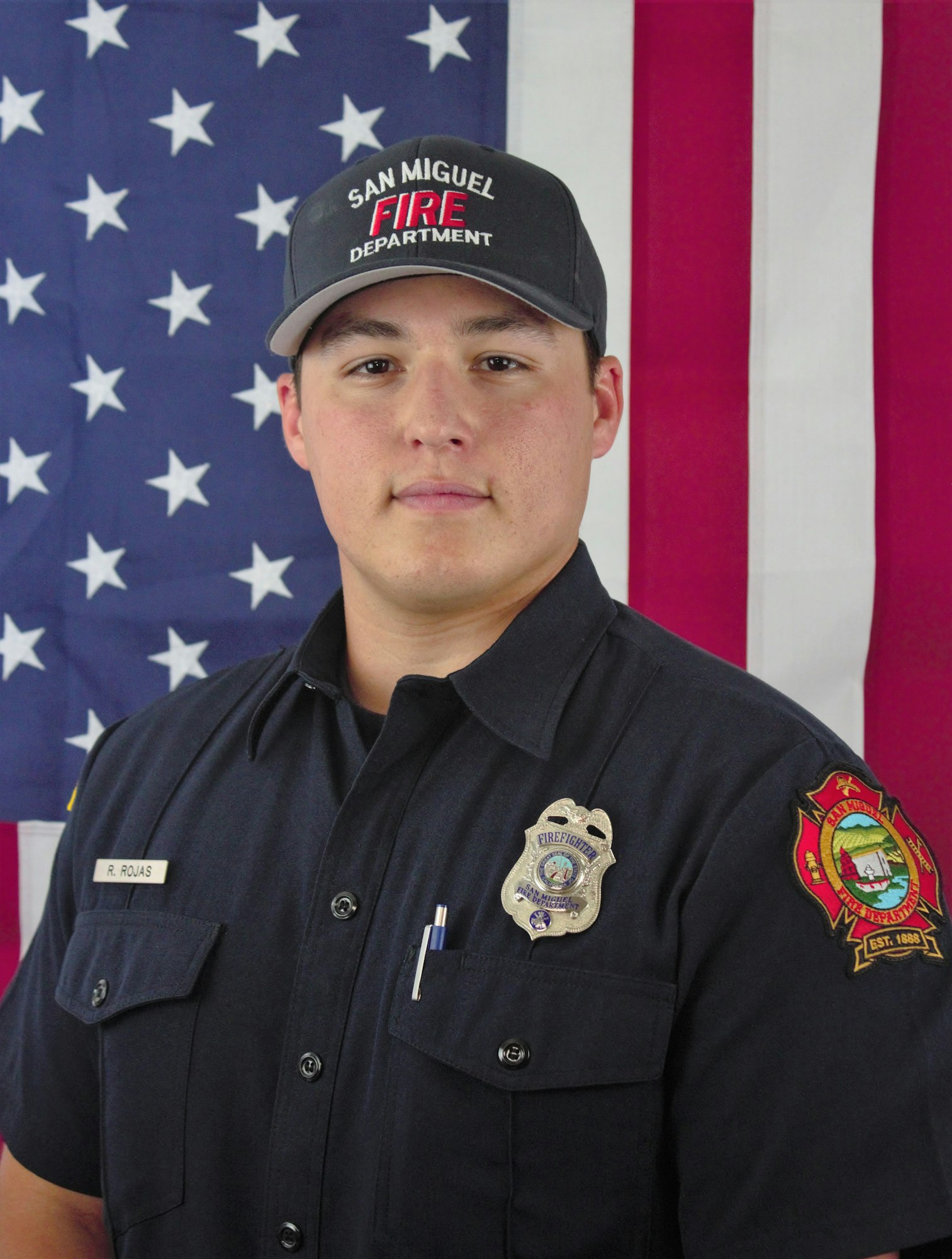 A firefighter in uniform stands in front of an American flag, wearing a hat with "San Miguel Fire Department" on it.