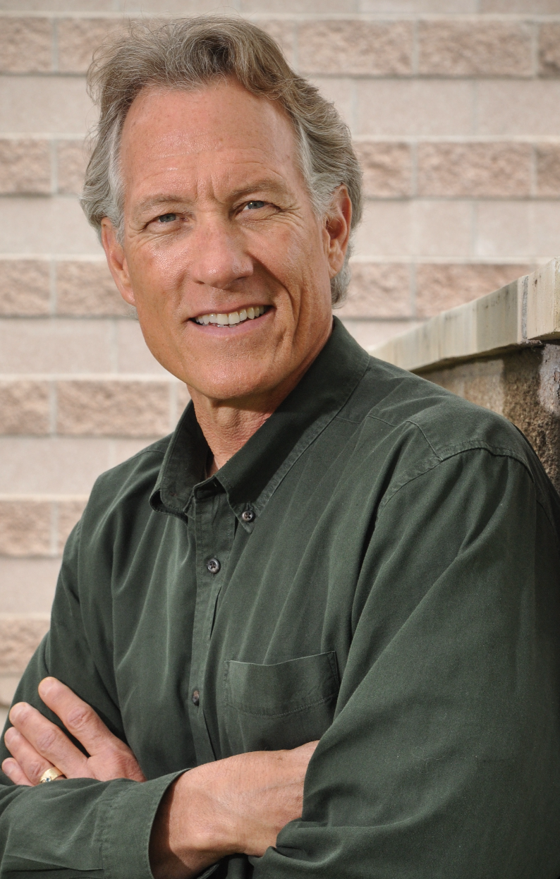 A smiling man with gray hair, in a green shirt, leaning against a brick wall with arms crossed.