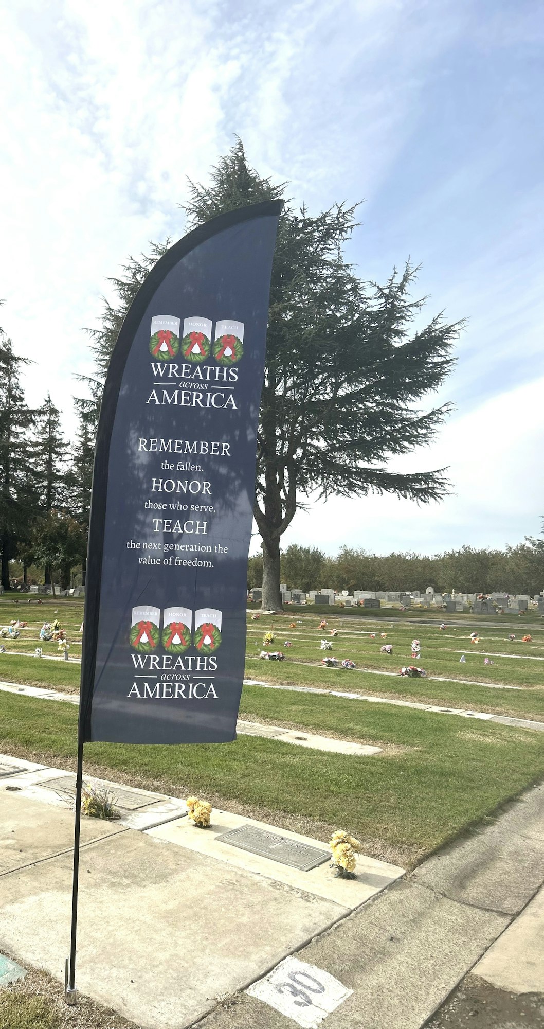 Banner for "Wreaths Across America" at cemetery with headstones and a tree in the background.