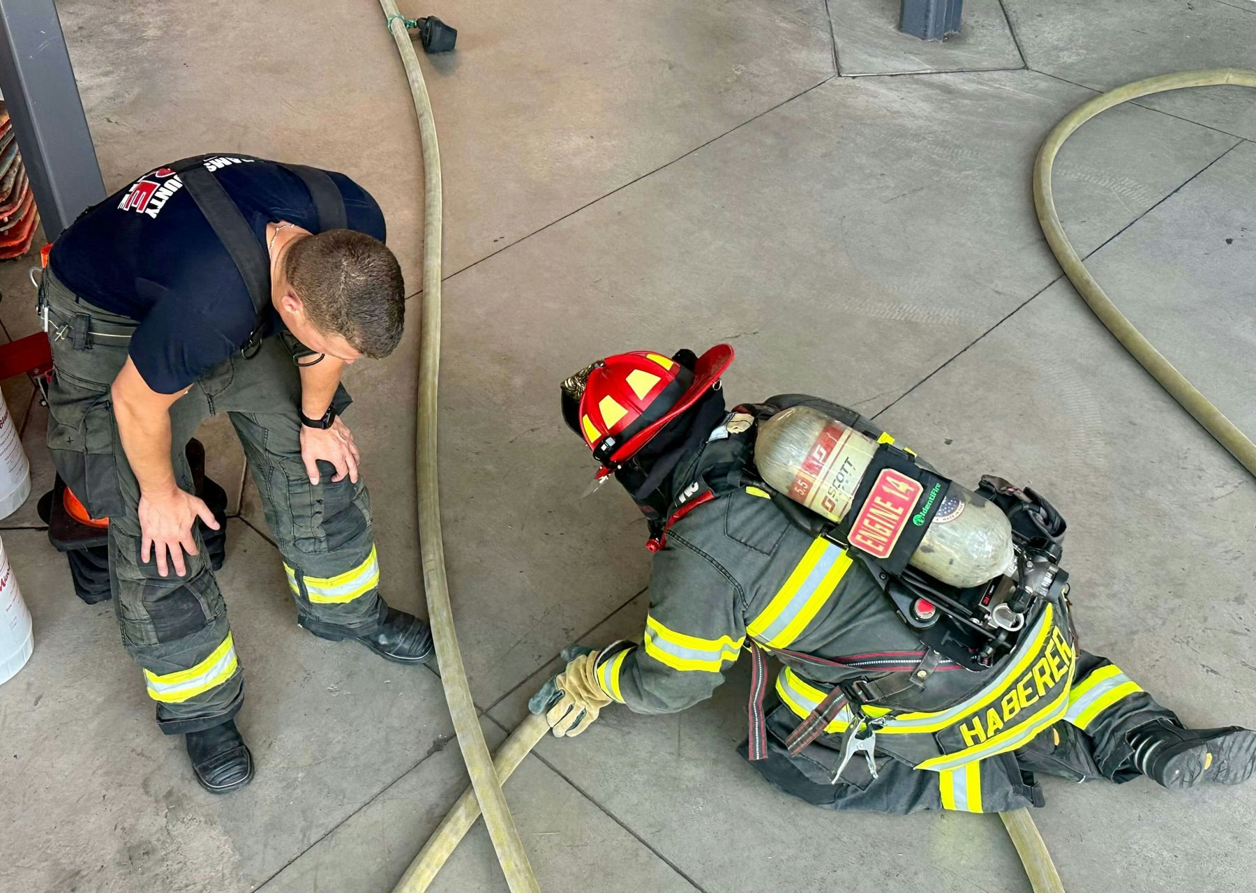 Two firefighters in gear, one crouching and the other kneeling, handling a fire hose in a station.