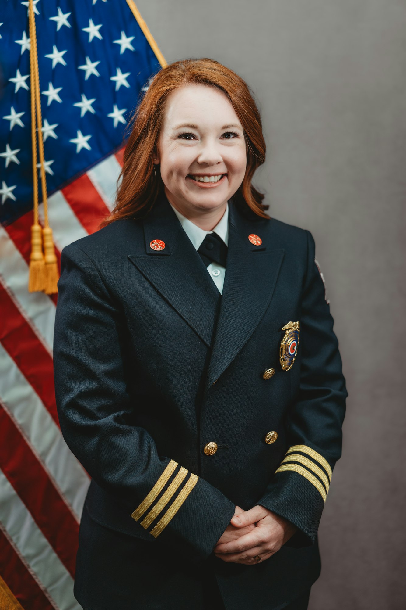 A person in a dark uniform stands smiling in front of an American flag.