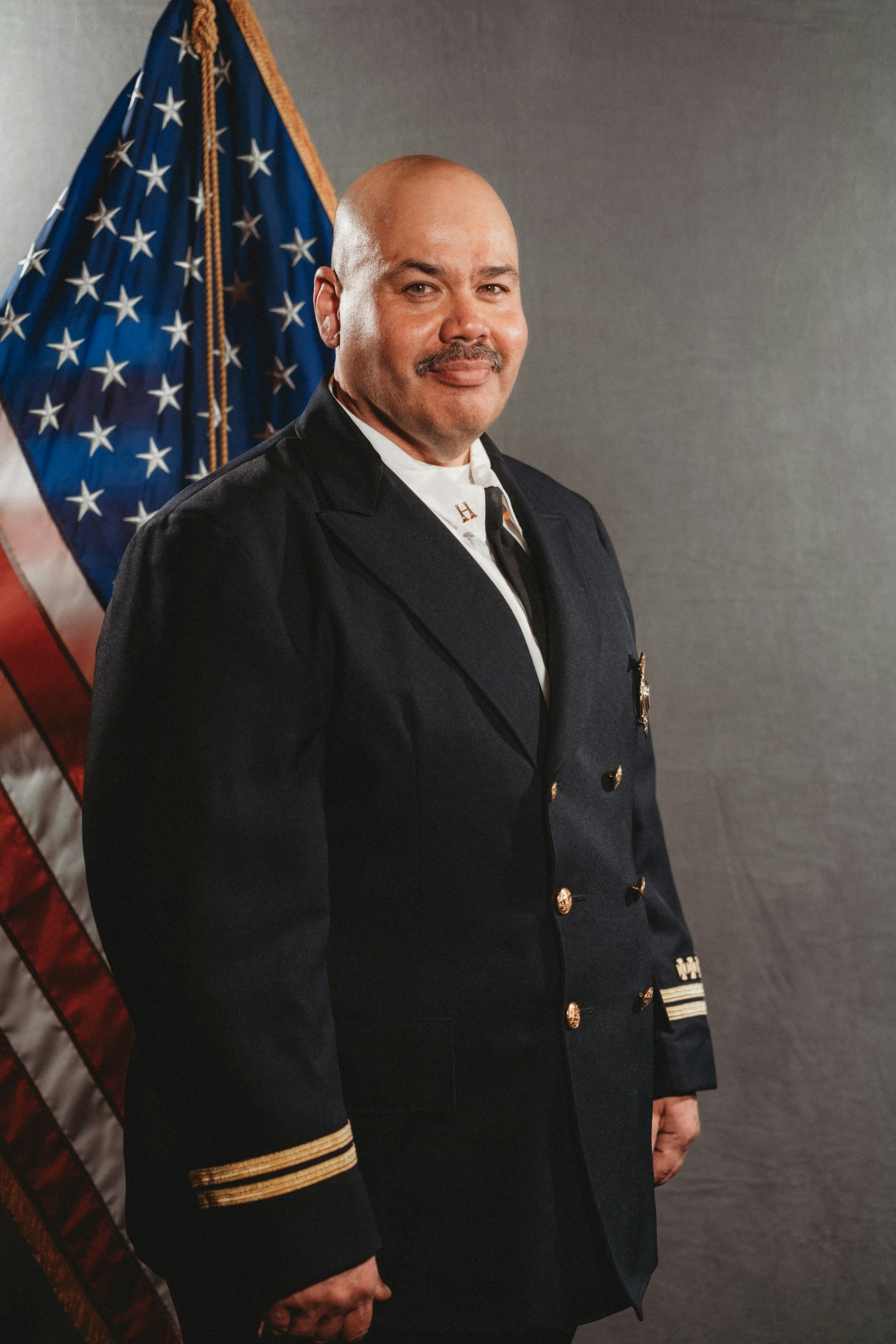 A man in uniform stands and smiles in front of a U.S. flag.
