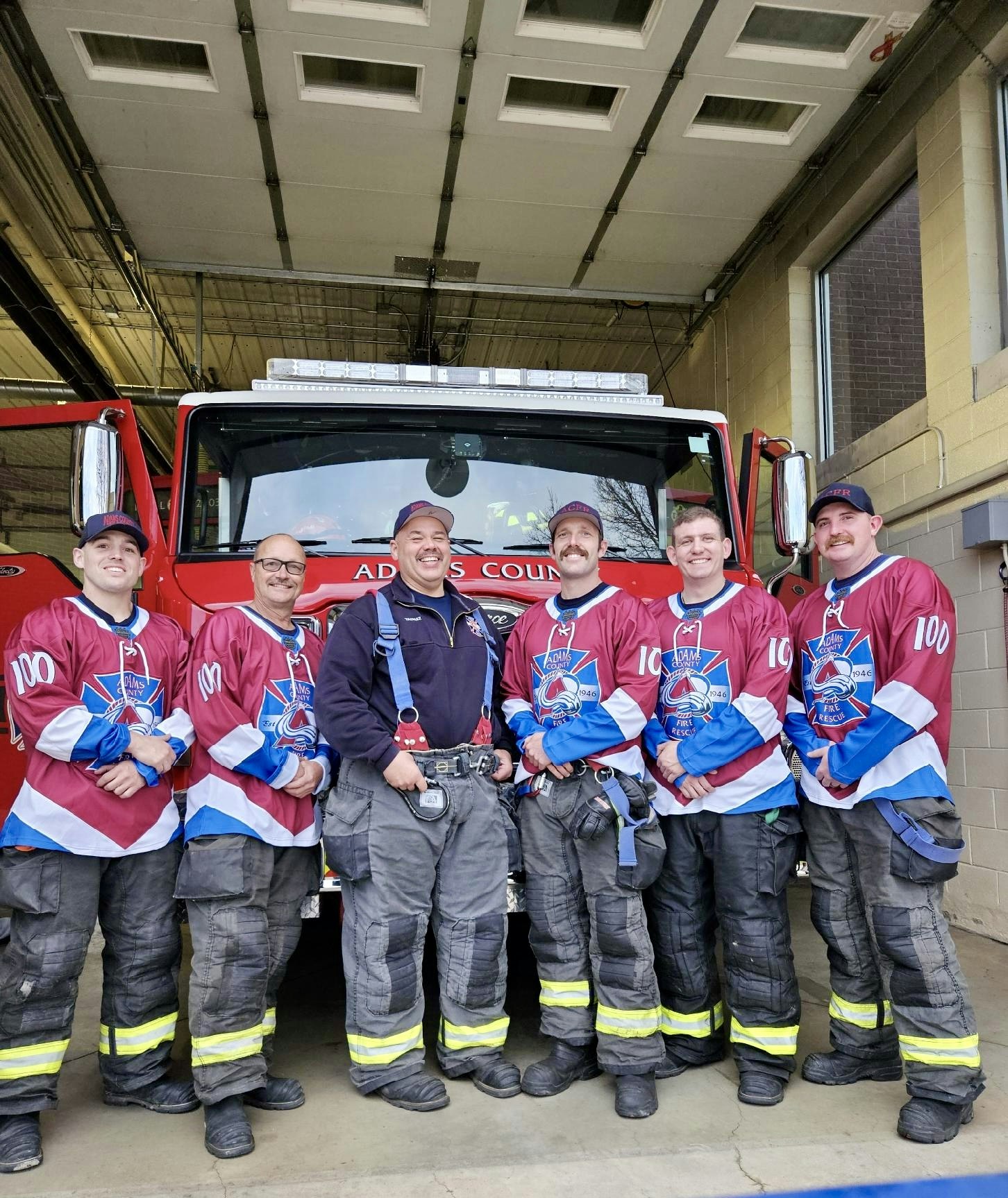 Six firefighters in jerseys posing in front of a fire truck inside a station.