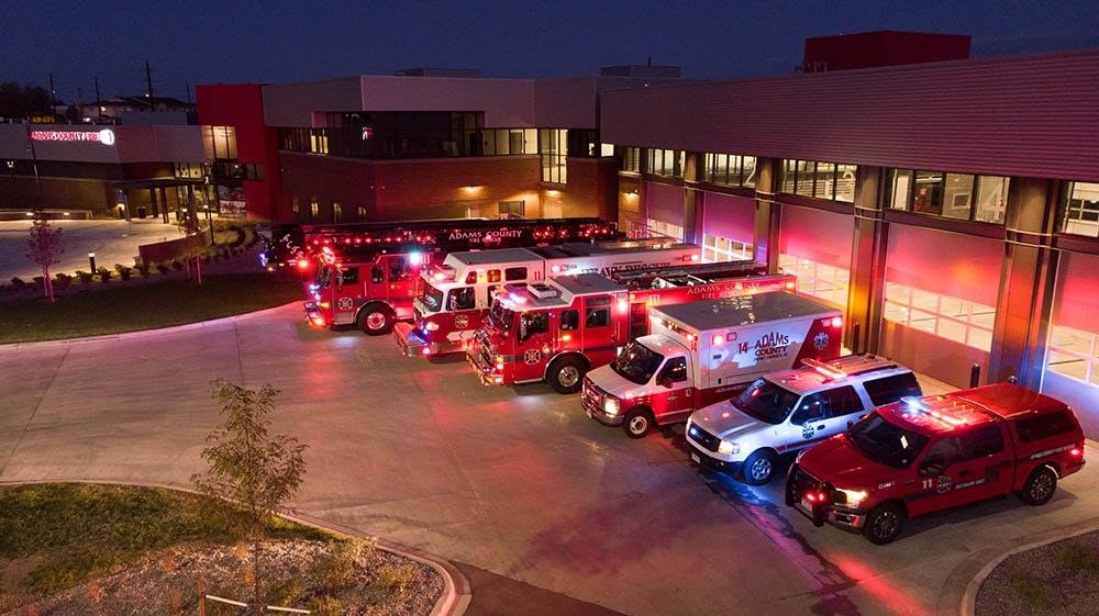 Fire trucks and emergency vehicles parked outside a fire station at night with lights on.