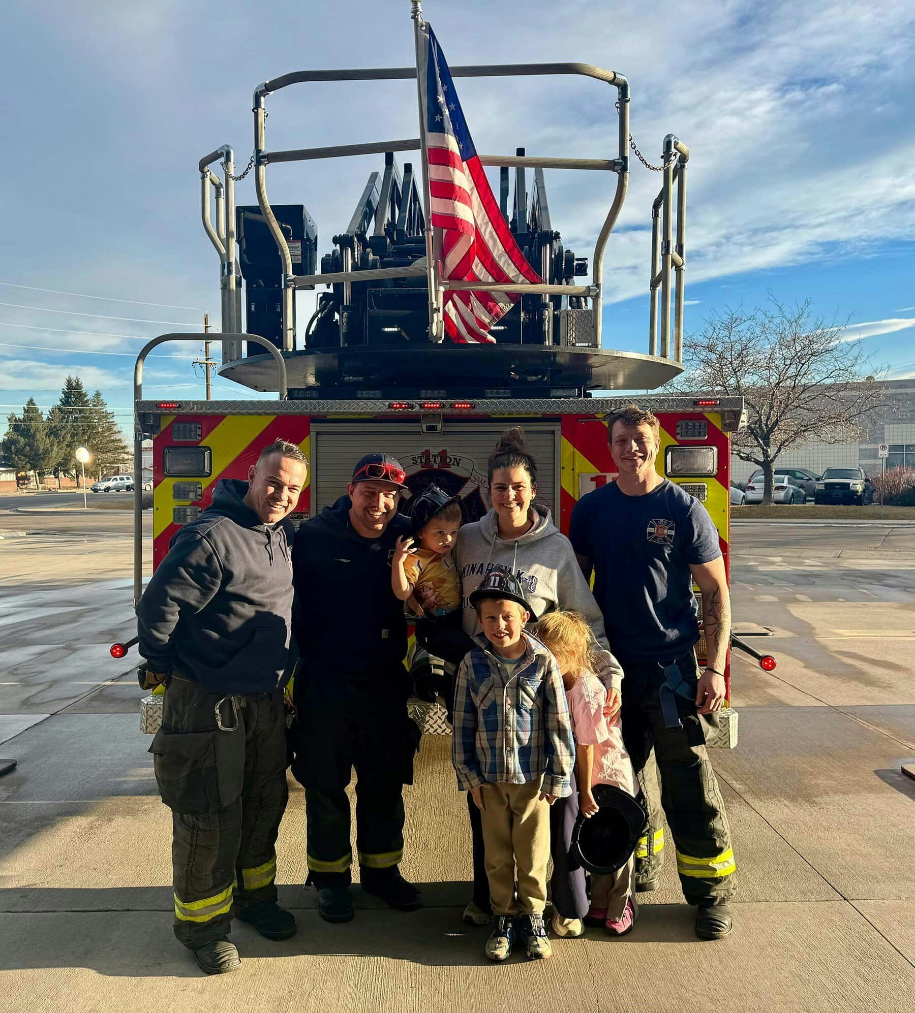 Five people and three children pose smiling in front of a fire truck with an American flag.