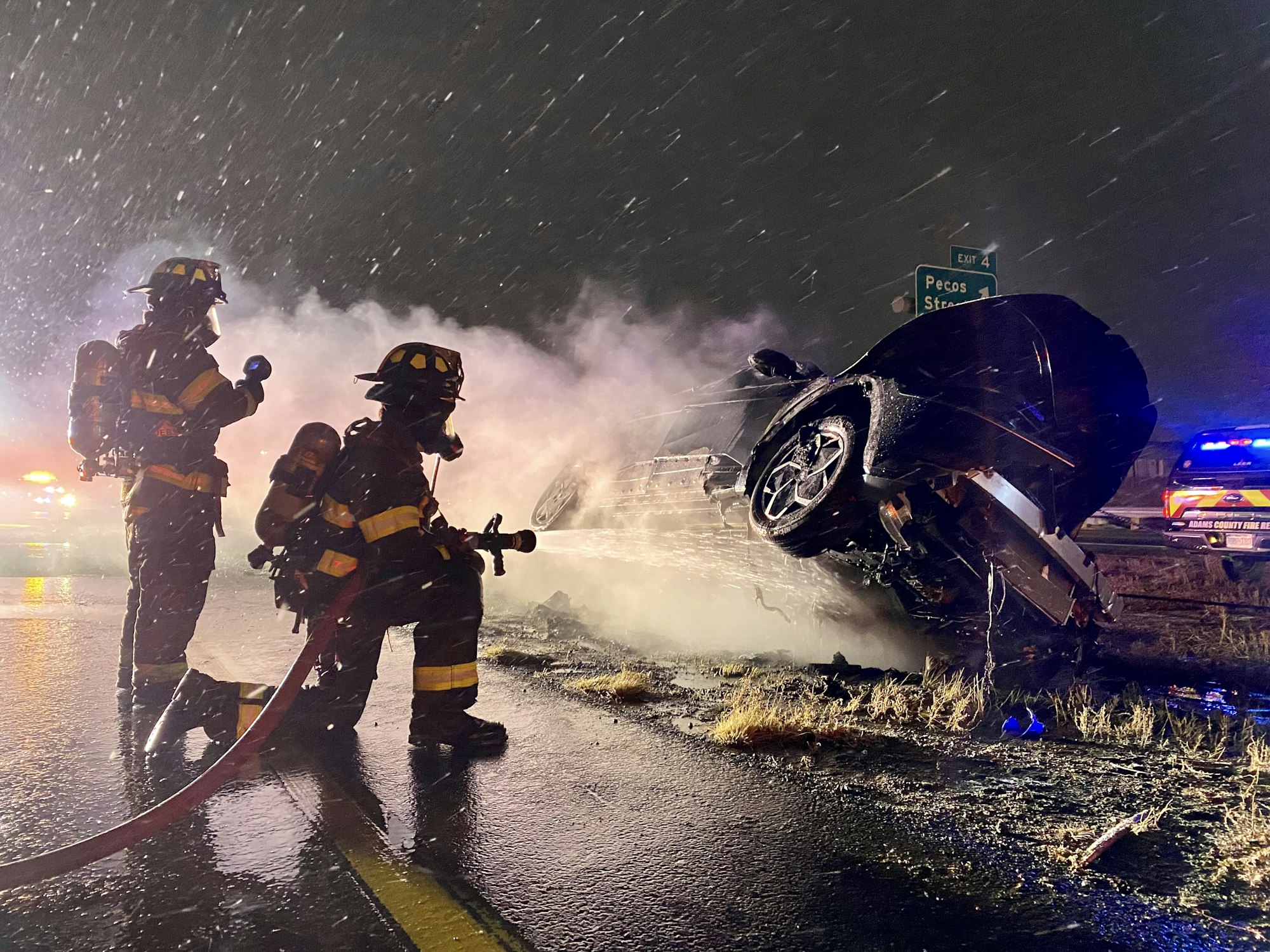 Firefighters respond to a nighttime car accident with an overturned vehicle, surrounded by smoke and emergency lights.