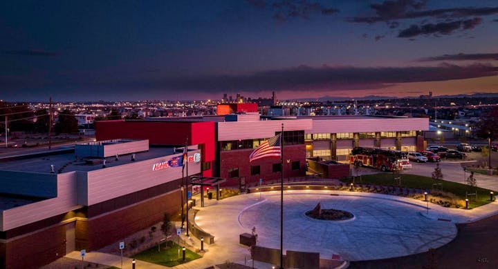 Fire station at dusk with an American flag, fire trucks, and city skyline in the background.