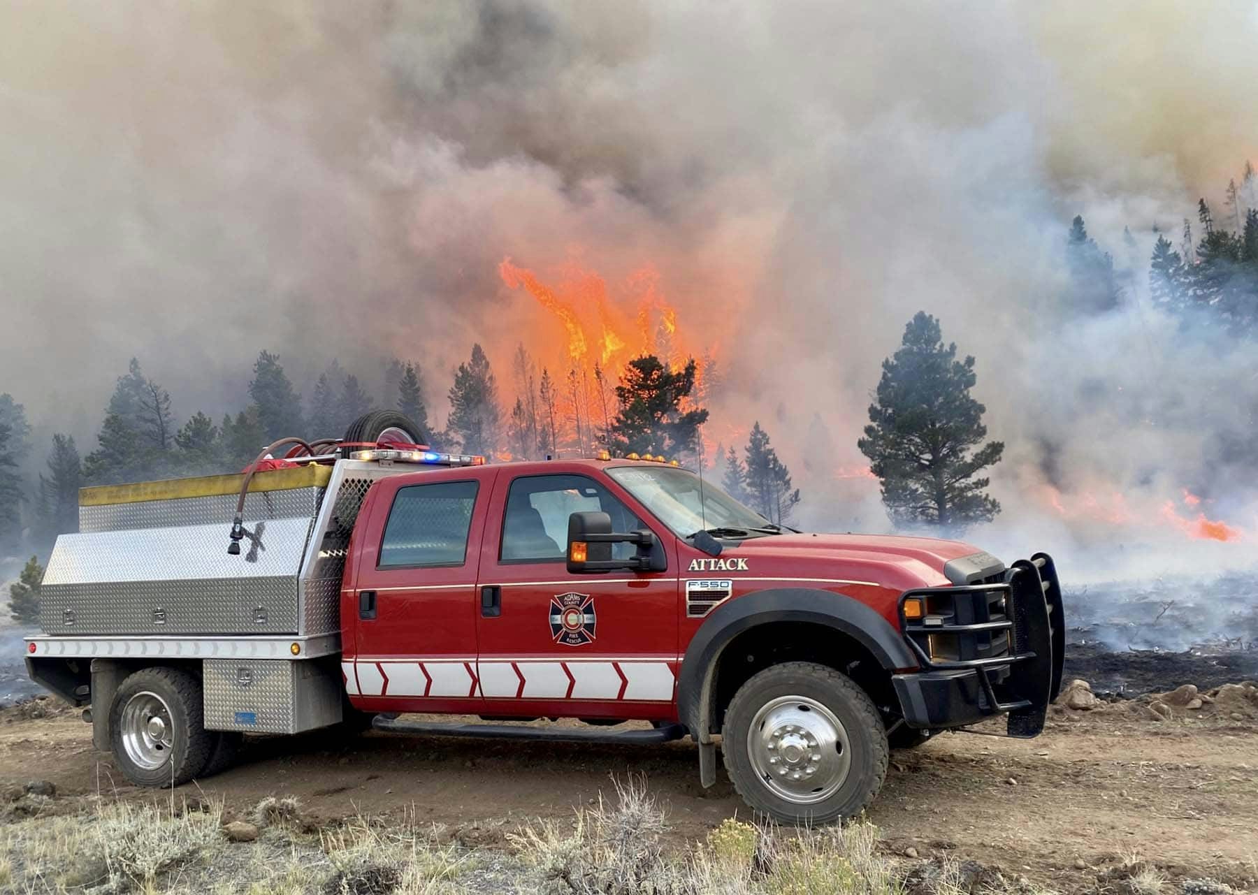 A red fire truck is near a forest fire with large flames and thick smoke.