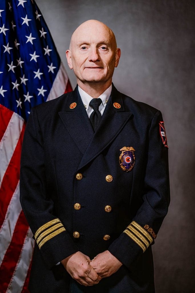 A uniformed official stands in front of a U.S. flag.