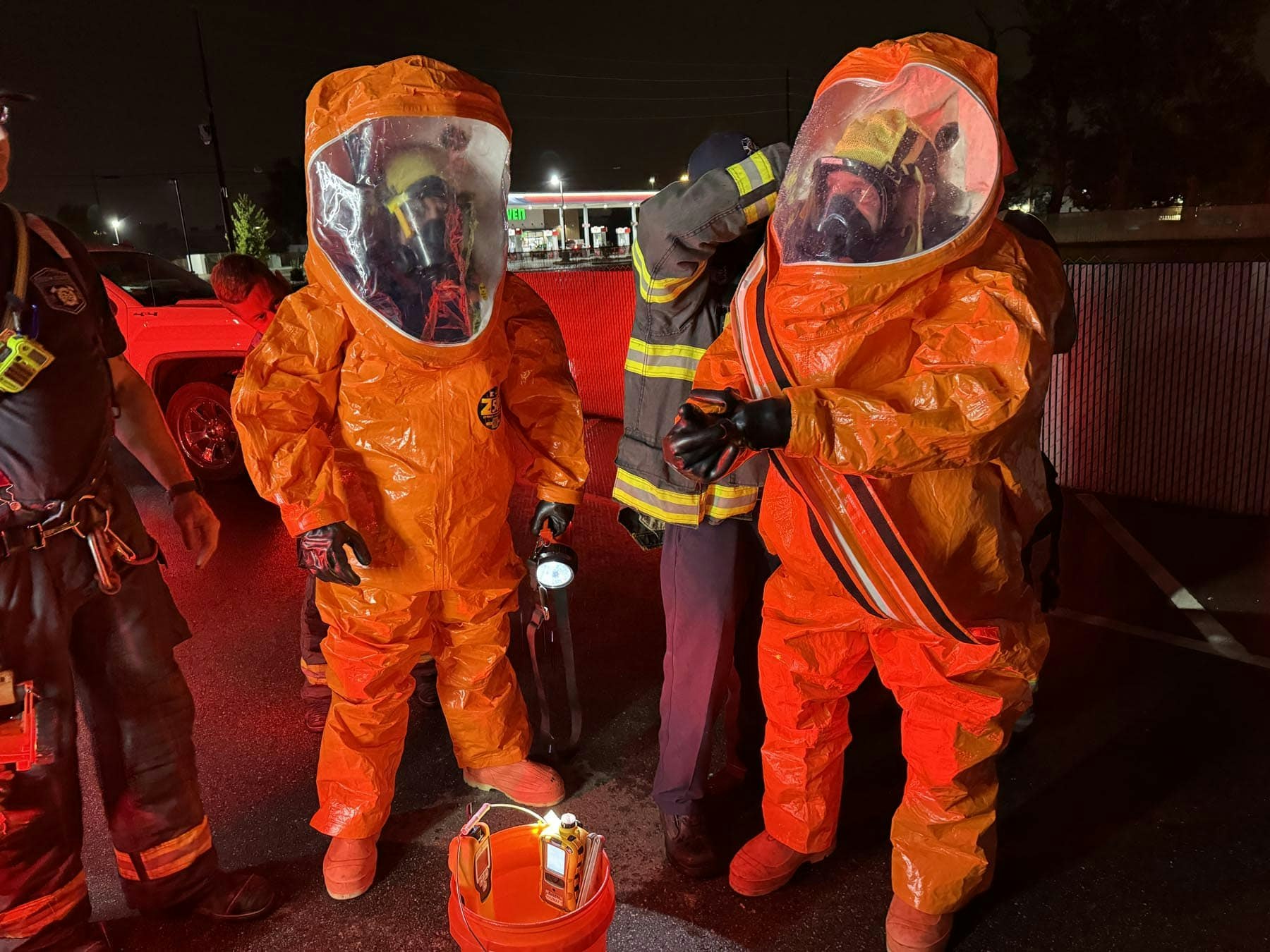 Two people in hazmat suits being assisted by firefighters under red lighting, holding a flashlight and standing near a bucket.