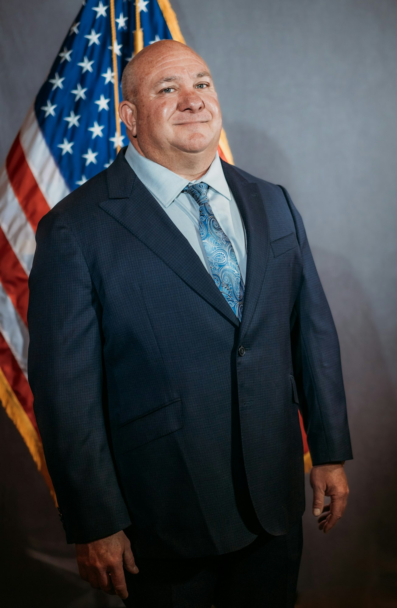 A man in a suit stands in front of a U.S. flag with a neutral background.