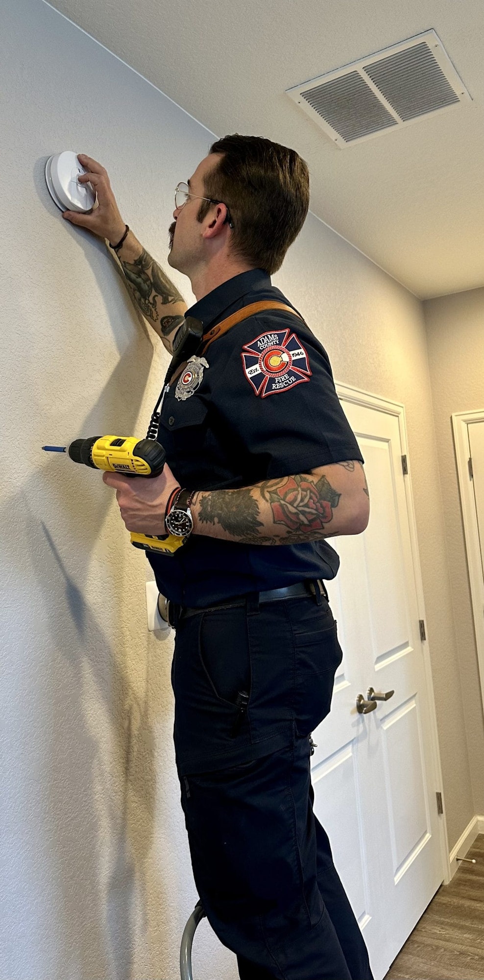 A firefighter on a stepladder installs a smoke detector on a wall, holding a drill.