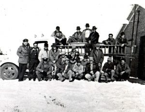 A group of men, some in uniform, gathered on and around a vintage fire truck in snowy surroundings.