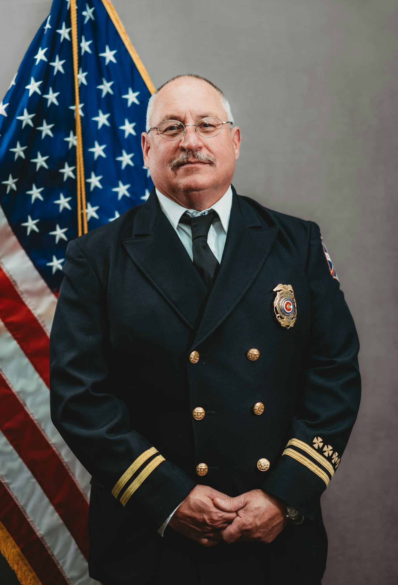 A man in a dark uniform with gold buttons and badges stands in front of an American flag.