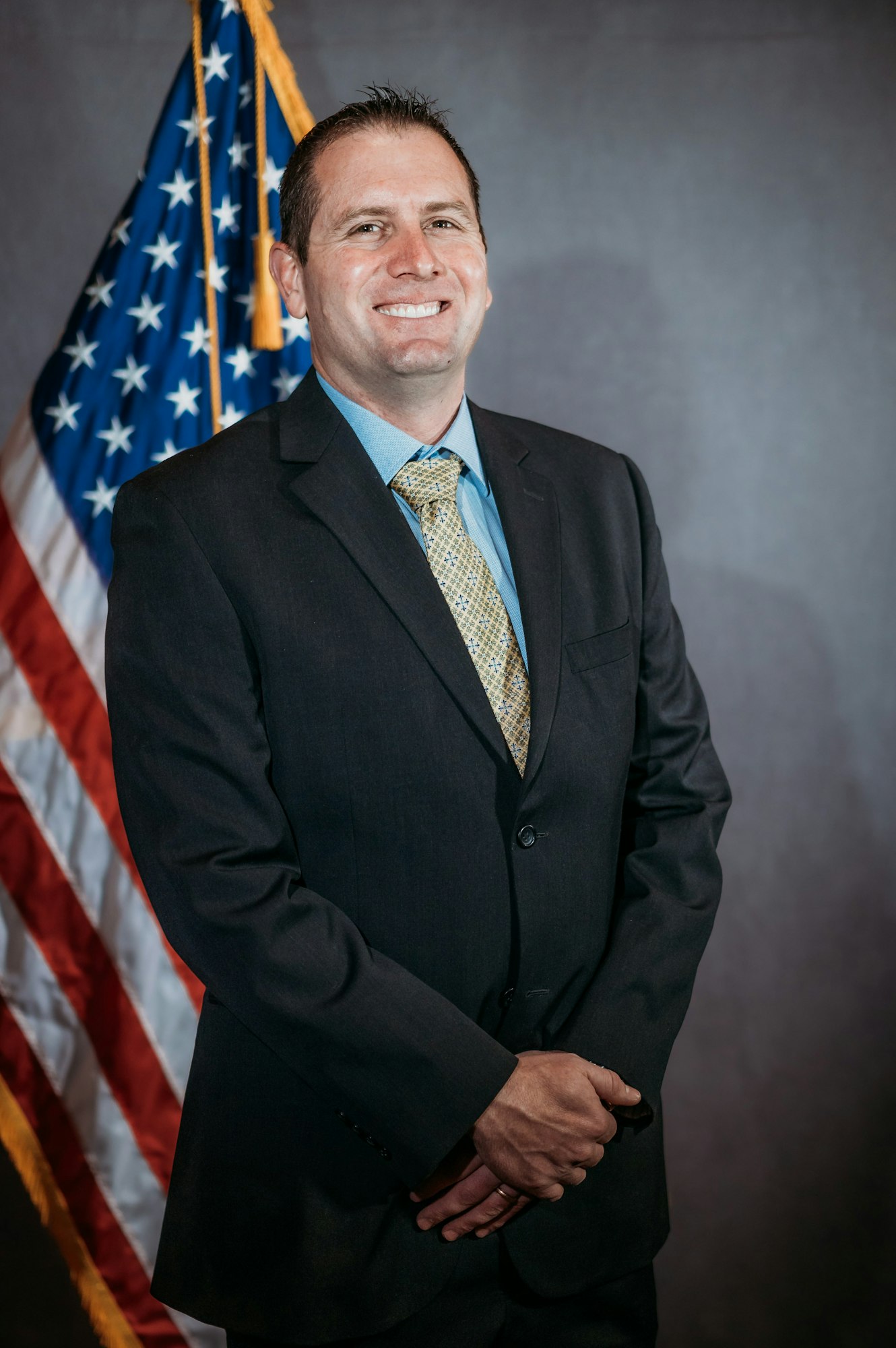 A man in a suit stands smiling in front of the U.S. flag.