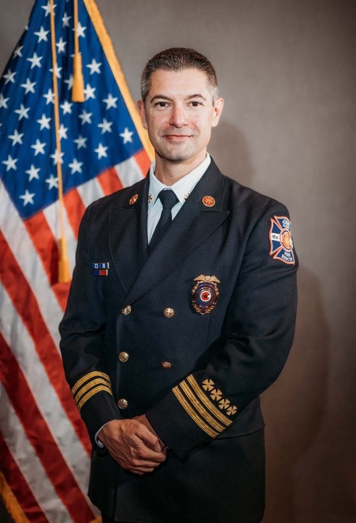 A person in a formal uniform stands in front of a U.S. flag.
