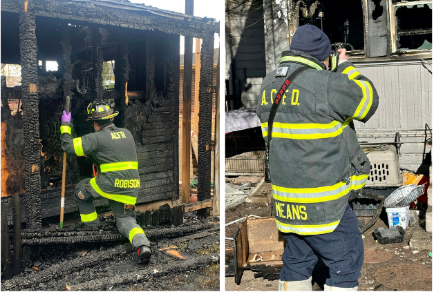 Two firefighters in gear inspect a charred structure, one examining debris and the other taking photographs.
