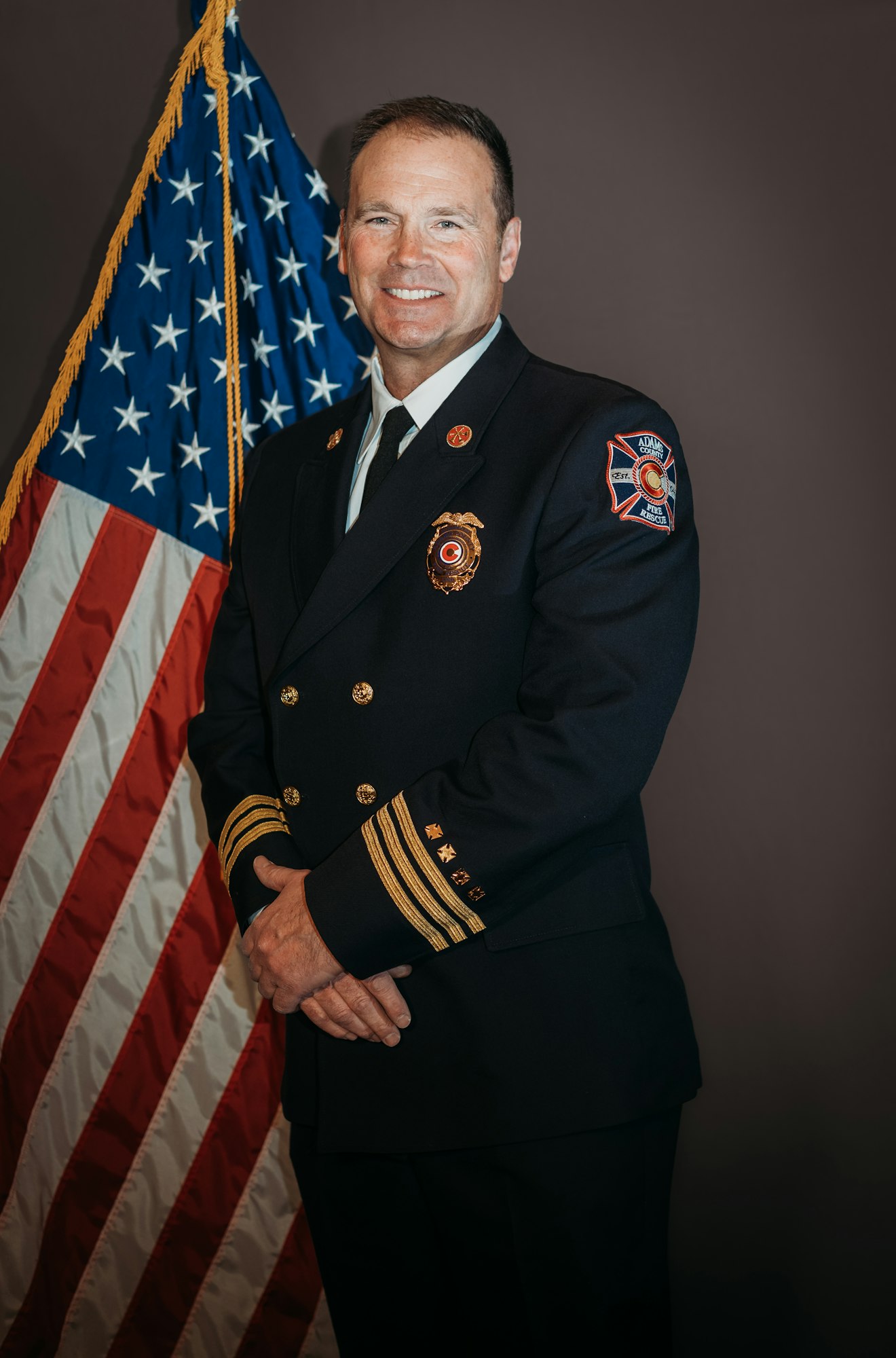A person in a formal uniform stands in front of an American flag, smiling.