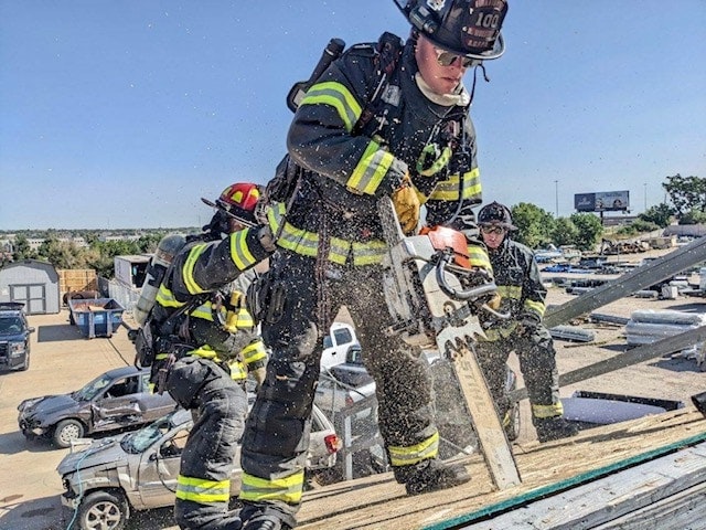 Firefighters cutting a roof with a chainsaw, sawdust flying, wearing full gear and helmets outdoors.