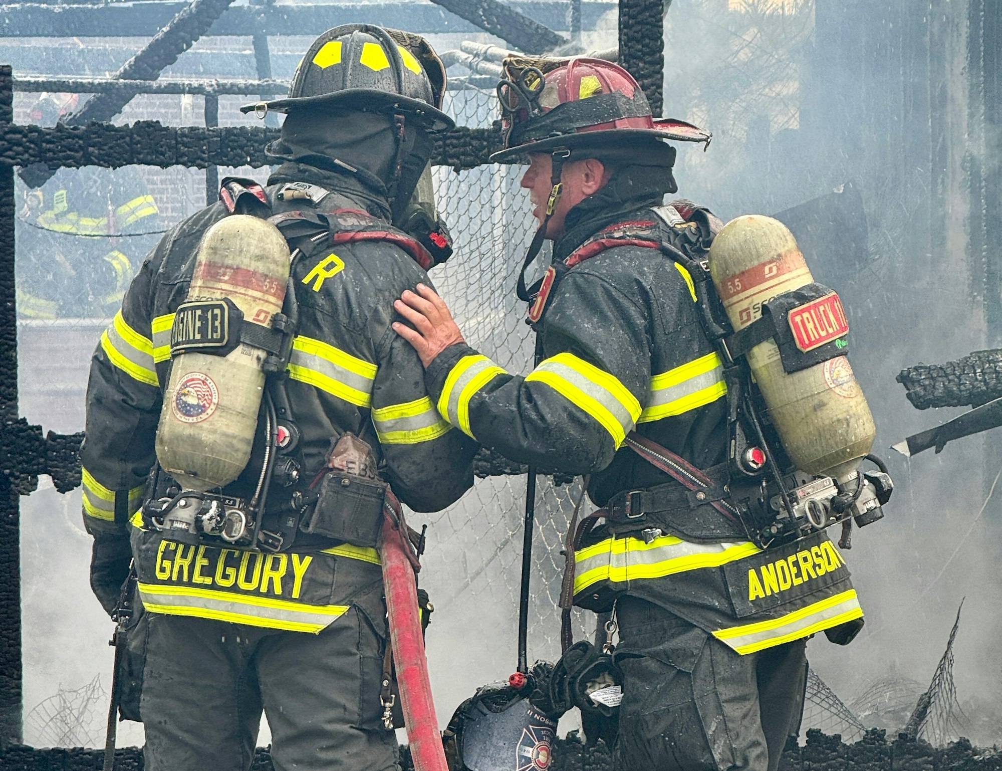 Two firefighters converse in smoky conditions, wearing protective gear and respirators, amidst a fire scene.